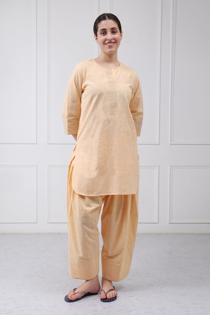 A woman modeling a light peach-beige three-piece kurta set with wide-leg pants and a matching tunic, standing against a white paneled wall.