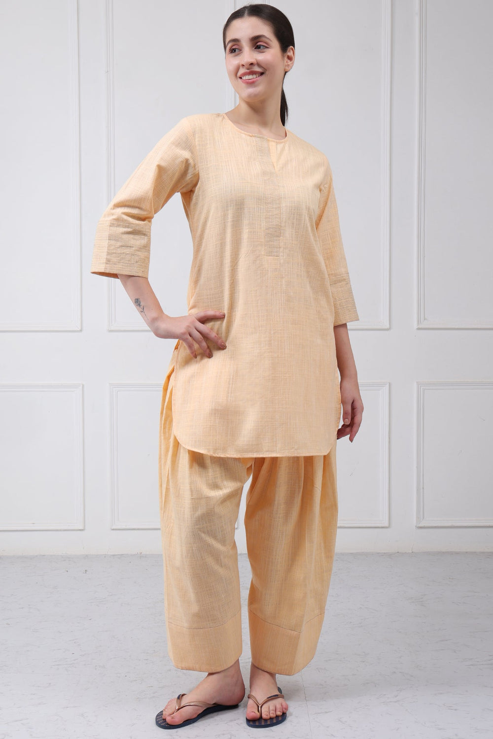 A woman modeling a light peach-beige two-piece kurta and pant set against a white paneled wall.
