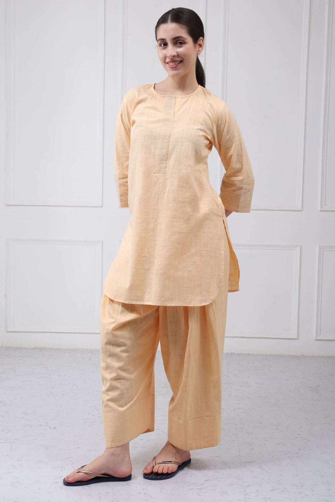 A woman modeling a light peach-beige two-piece traditional outfit consisting of a long-sleeve kurta and wide-leg pants, paired with black flip-flops, standing against a white paneled wall.