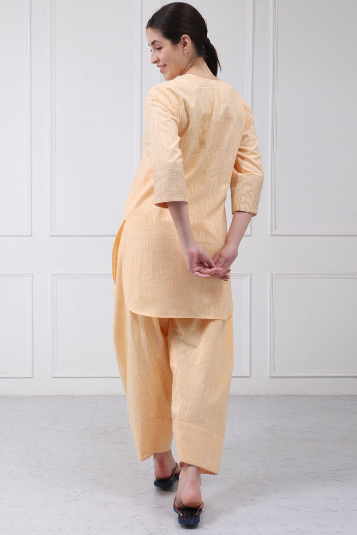 A woman wearing a light peach-colored linen kurta and wide-leg trousers set, shown from a back-three-quarter view against a white paneled wall.
