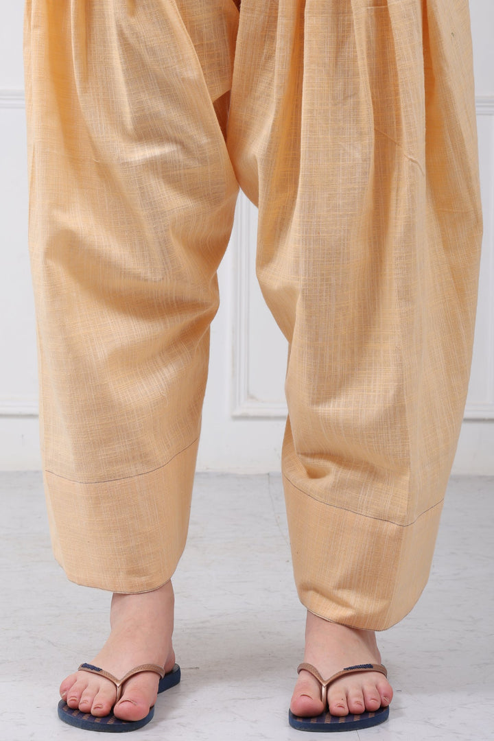 A person wearing loose, draped mustard-yellow or peach-colored harem-style pants with black thong sandals, shown from the waist down against a white paneled wall and light floor.