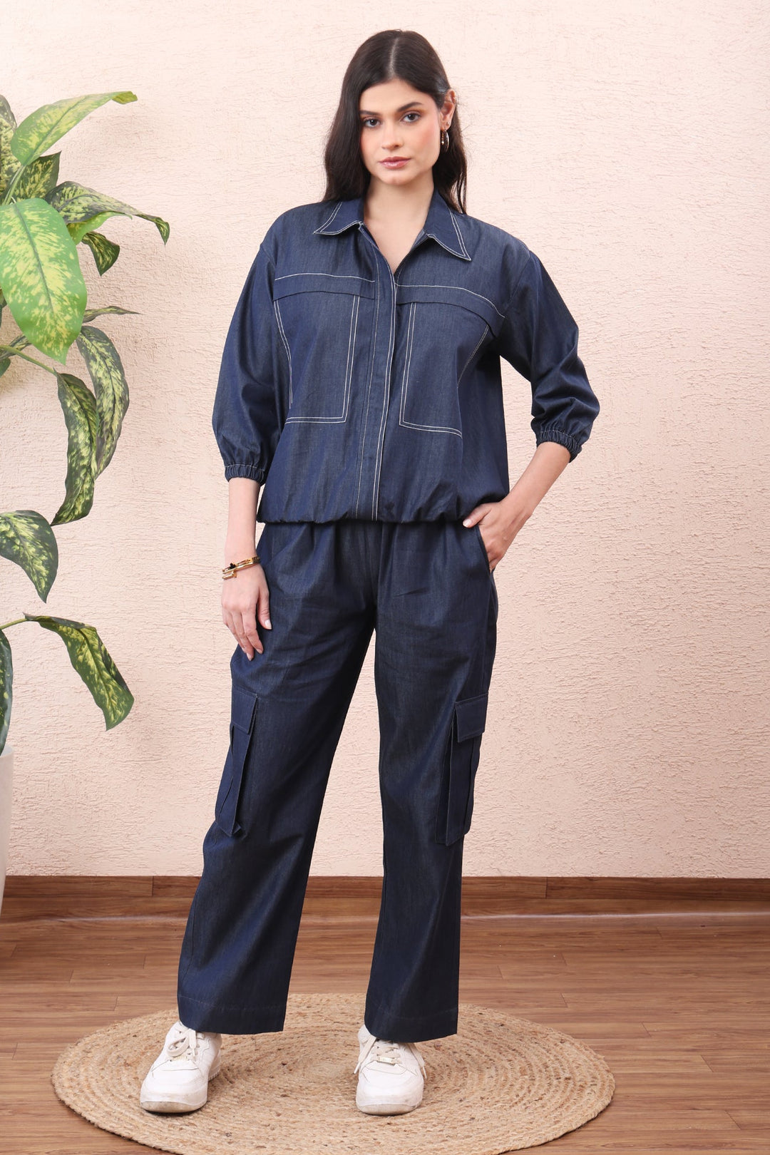 Woman wearing a blue jumpsuit standing in a room with a plant and wooden floor.