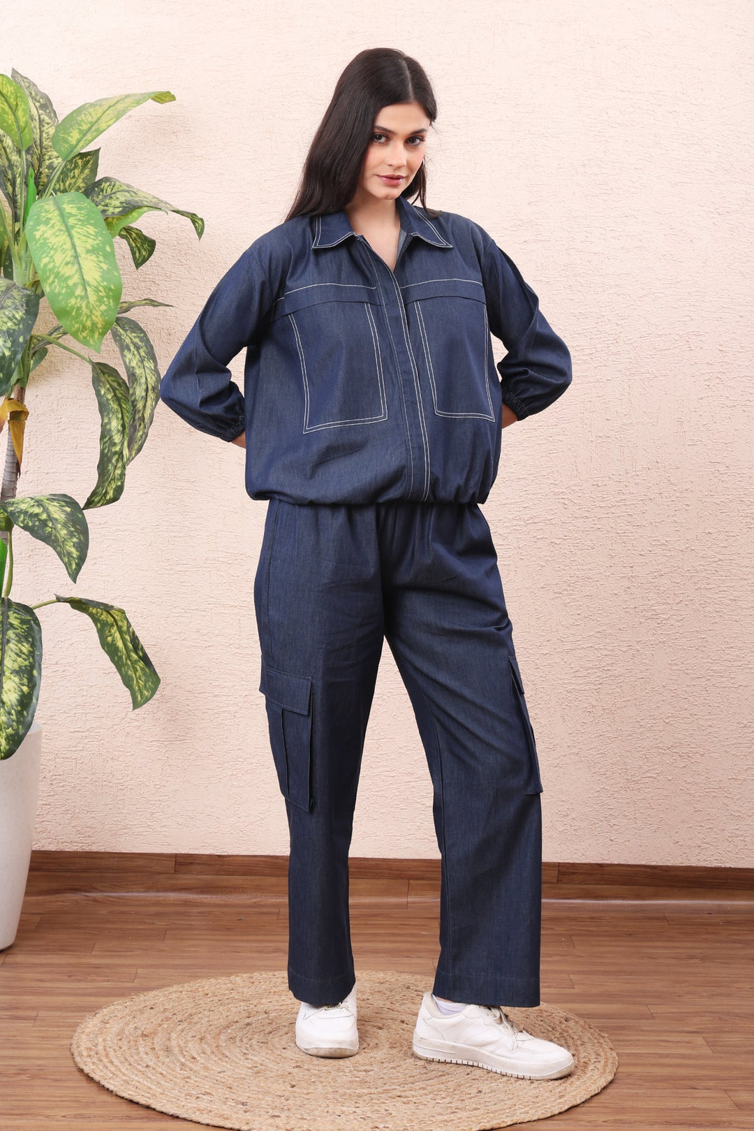 Woman wearing a navy blue jumpsuit standing in a room with a plant and wooden floor.