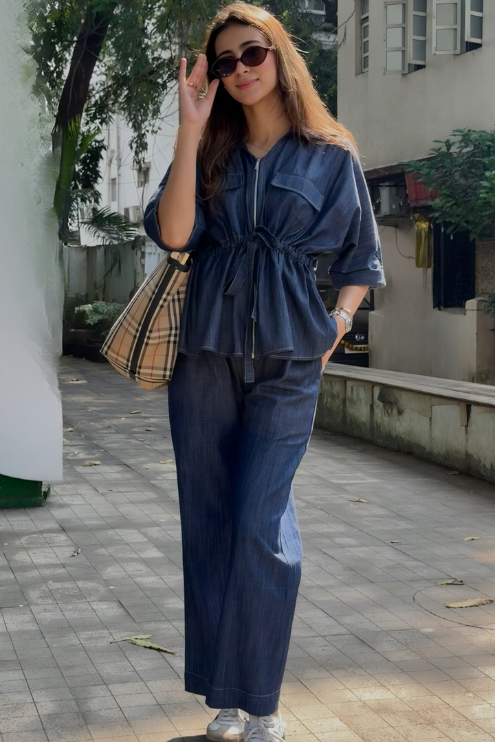 Woman in a denim outfit standing on a sidewalk with a building and trees in the background.