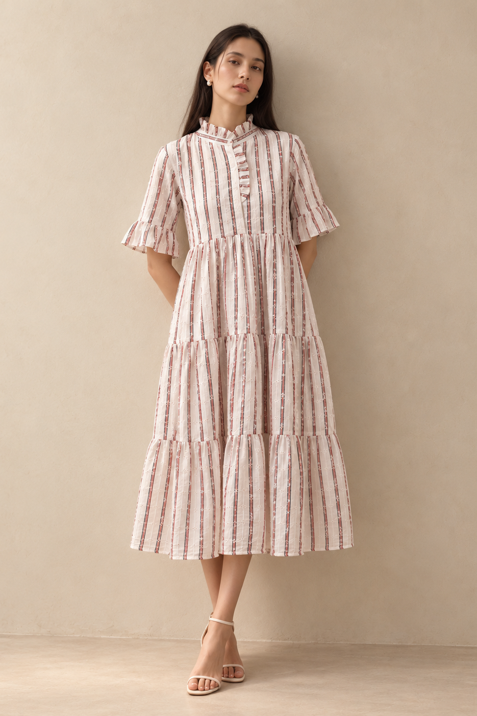 A woman stands against a plain beige wall wearing a tiered midi dress with vertical pink and white stripes and flutter sleeves, paired with light beige strappy heels.