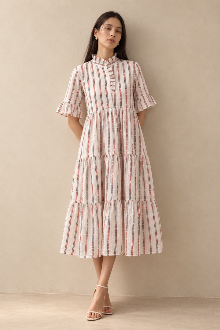 A woman stands against a plain beige wall wearing a tiered midi dress with vertical pink and white stripes and flutter sleeves, paired with light beige strappy heels.