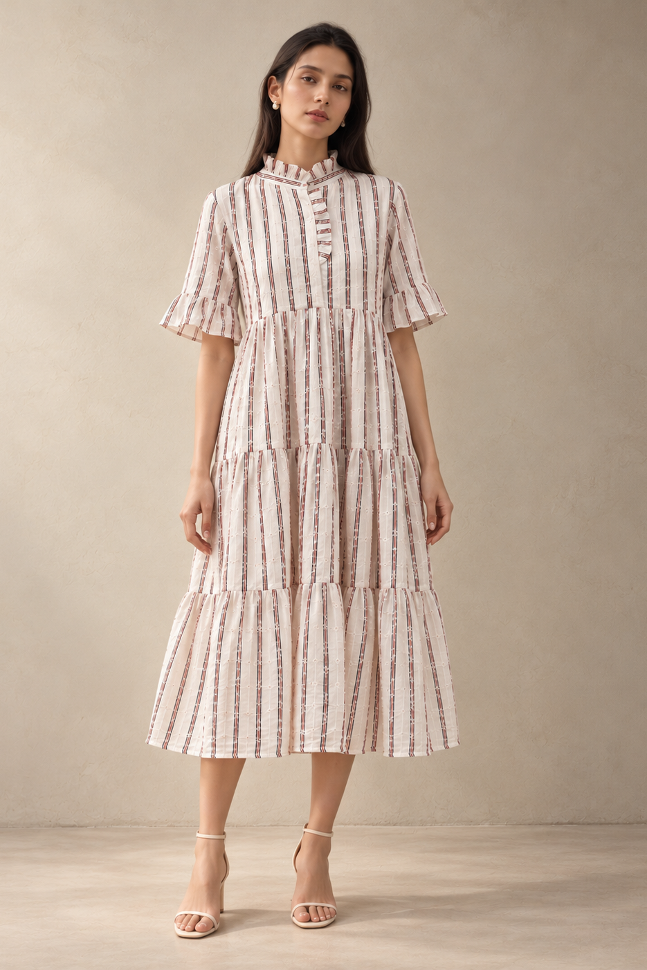 A woman modeling a tiered midi dress with vertical pink and beige stripes, ruffled short sleeves, and a high mock neck, standing against a neutral beige studio backdrop.