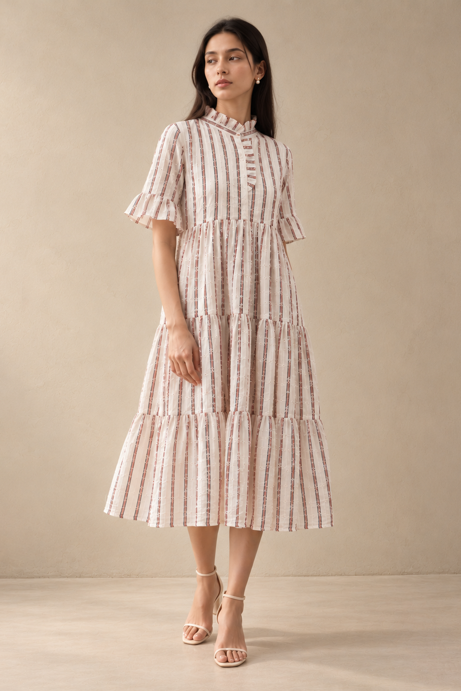 A woman models a tiered midi dress with vertical pink and beige stripes, featuring ruffled short sleeves and a high mock neck, paired with beige strappy heels against a neutral beige studio backdrop.