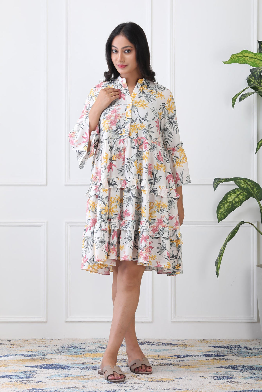 Woman wearing a floral dress standing in a room with a plant and white paneled wall.