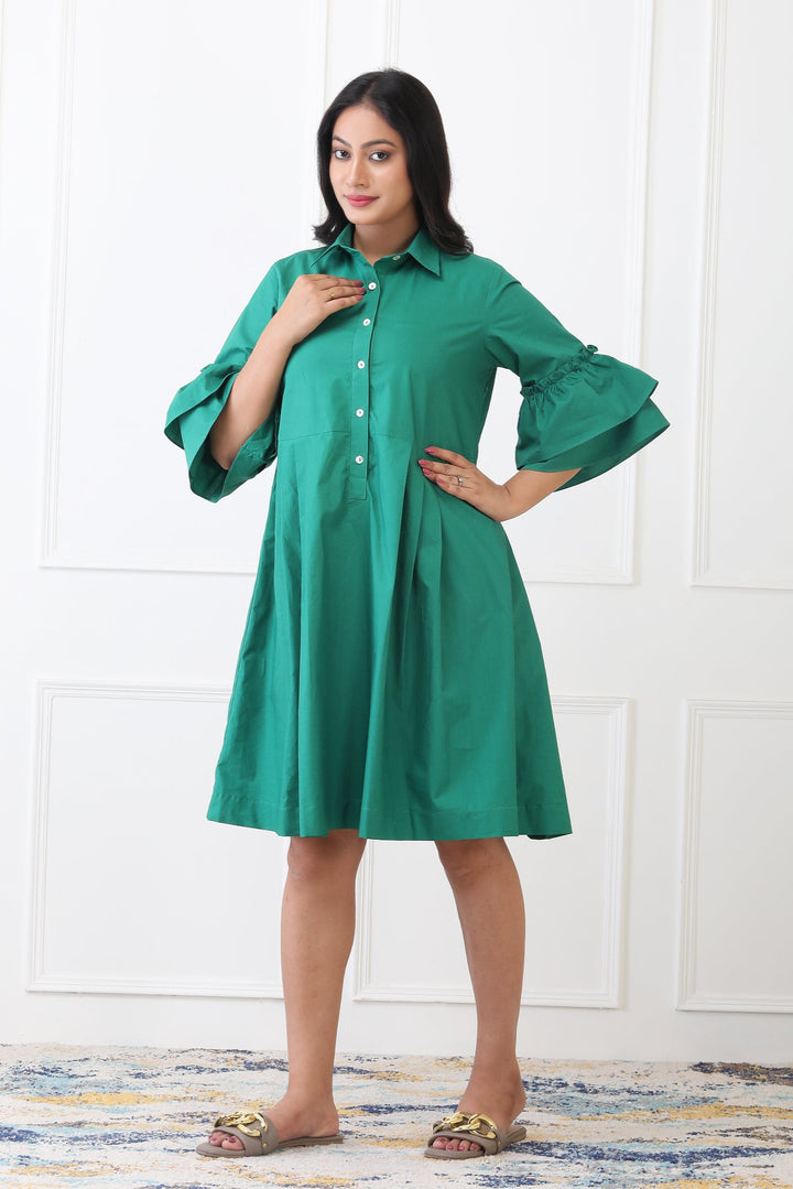 Woman wearing a green dress with ruffled sleeves against a white wall.