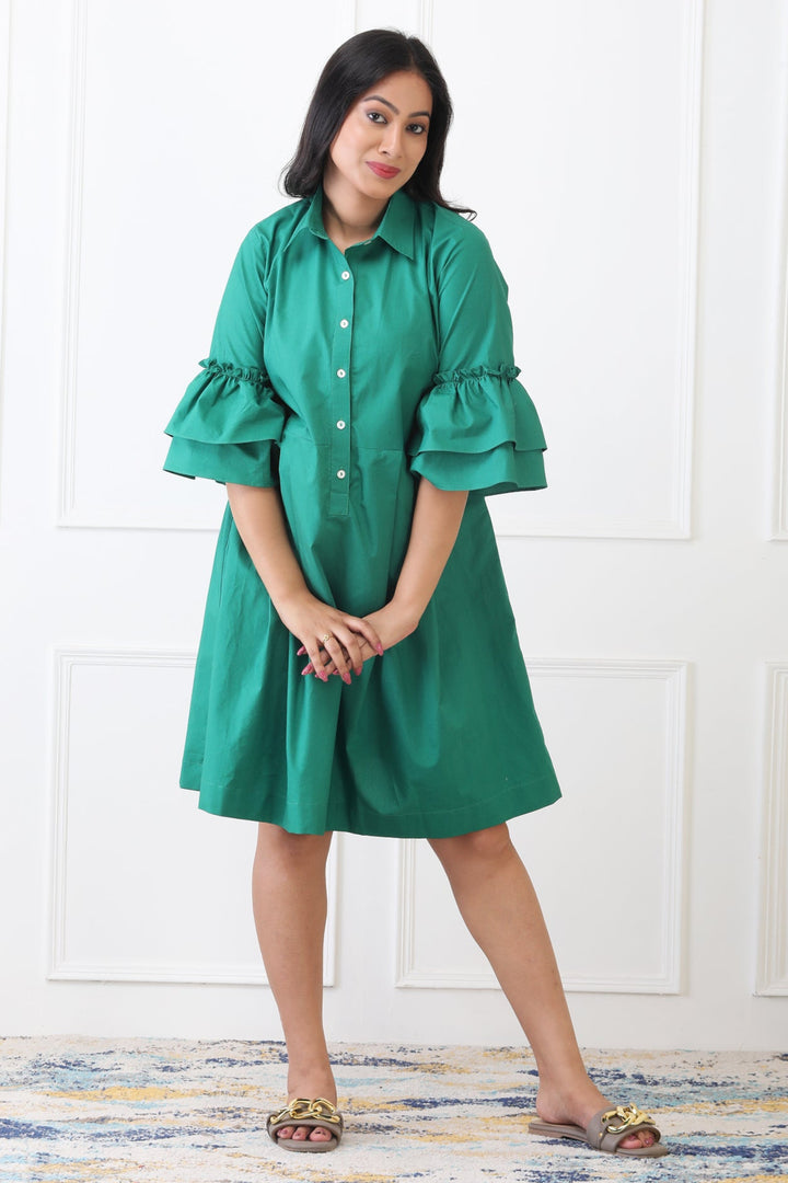 Woman wearing a green dress with ruffled sleeves against a white paneled wall.