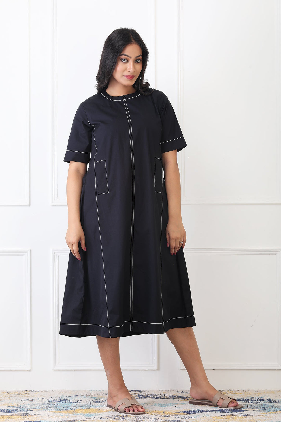 Woman wearing a black dress with white stitching against a white paneled wall.