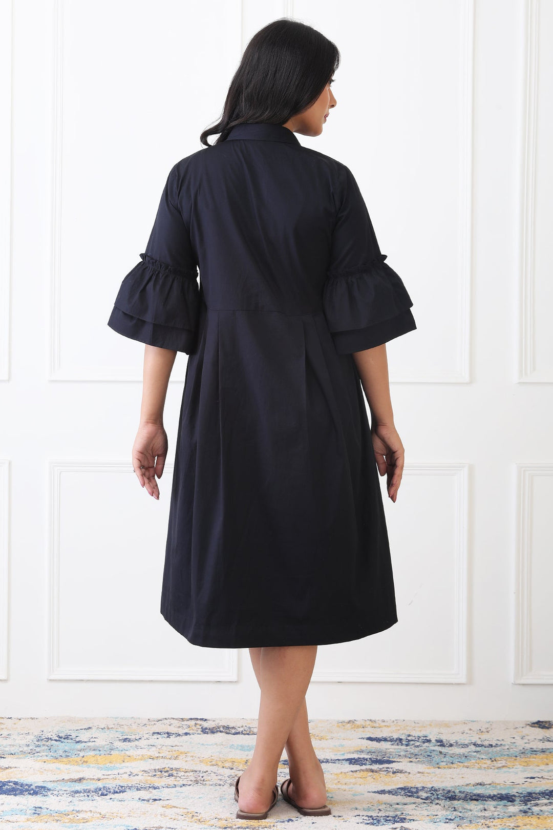 Woman wearing a black dress with ruffled sleeves against a white wall.