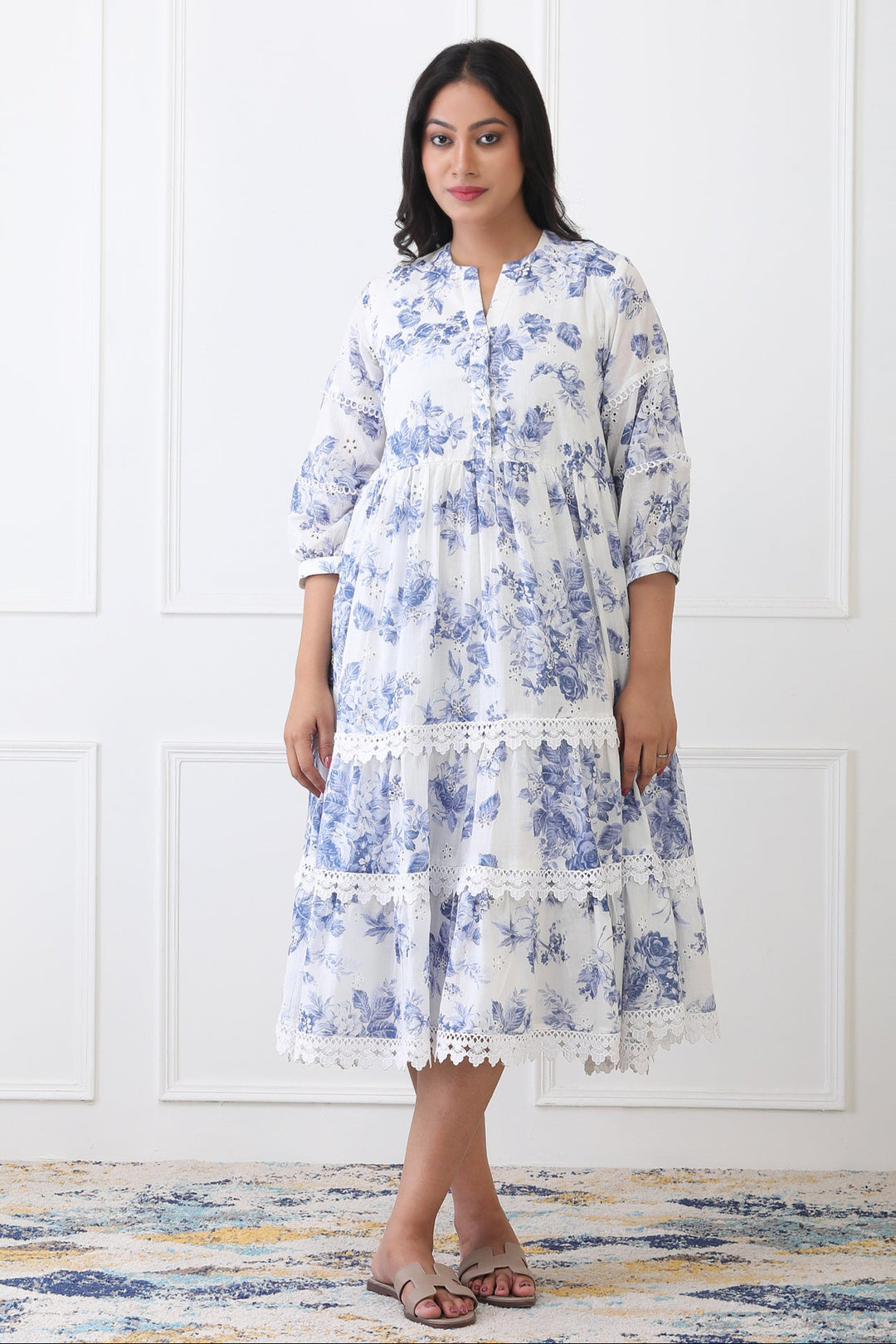 Woman wearing a blue floral dress standing against a white wall.