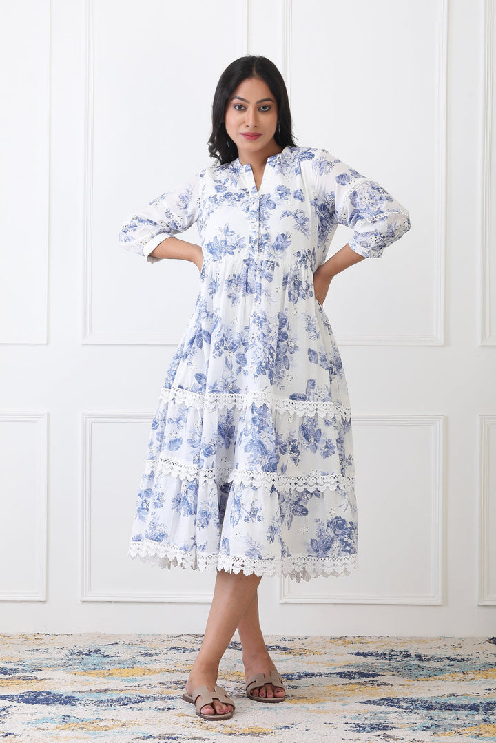 Woman wearing a blue floral dress standing against a white wall.