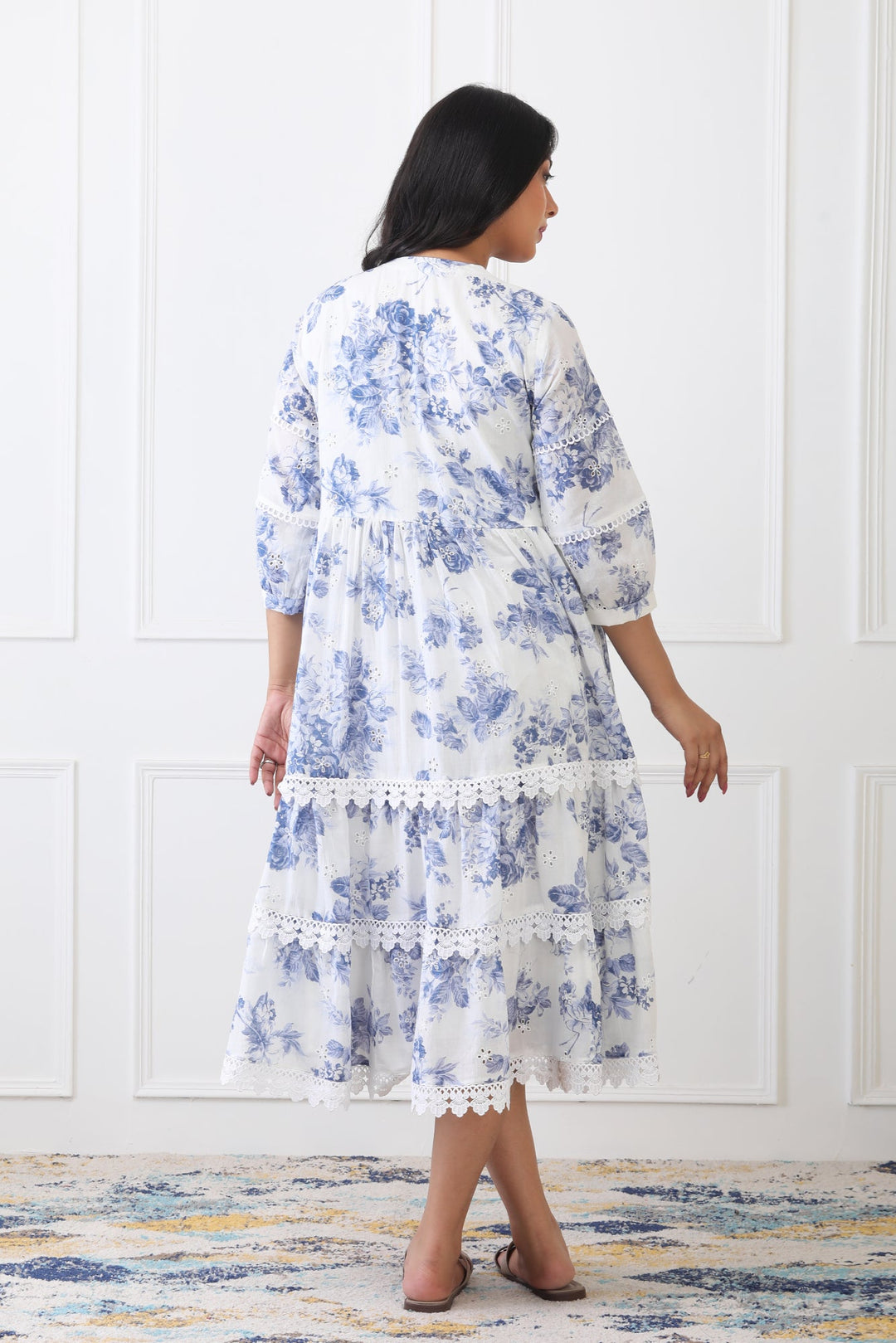 Woman wearing a blue floral dress standing against a white paneled wall.
