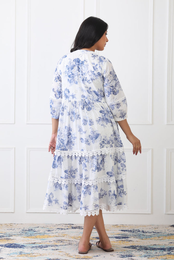Woman wearing a blue floral dress standing against a white paneled wall.