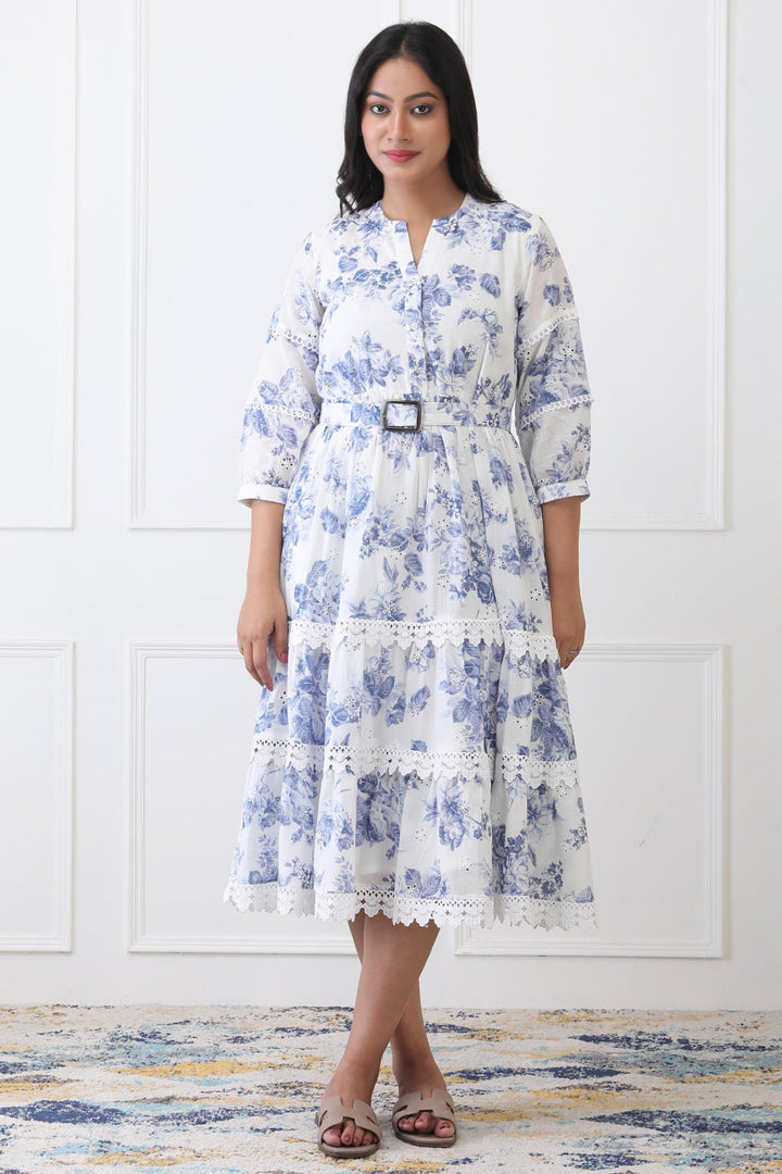 Woman wearing a white dress with blue floral pattern in a room with a white paneled wall.