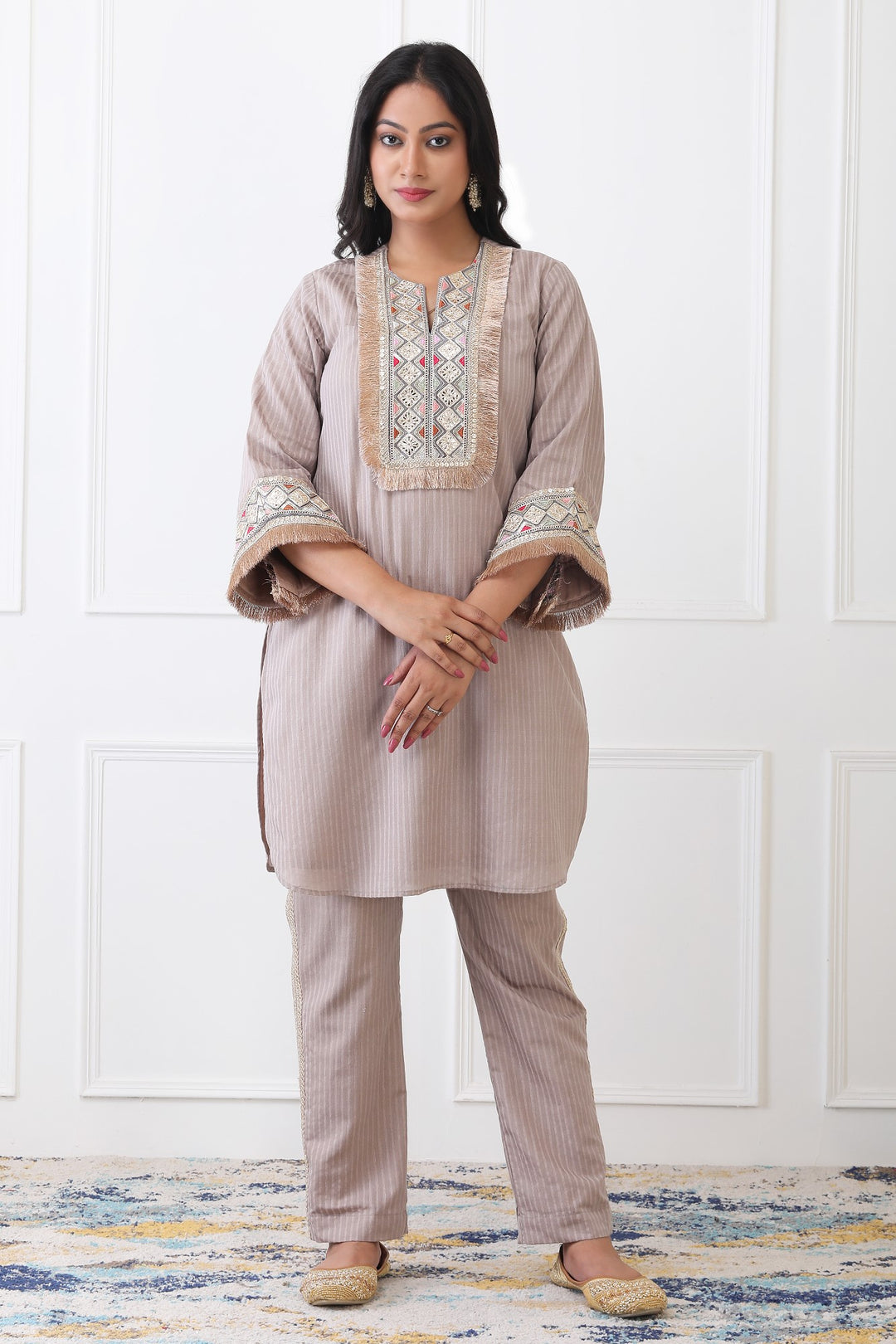 Woman wearing a beige traditional outfit with embroidery against a white paneled wall.