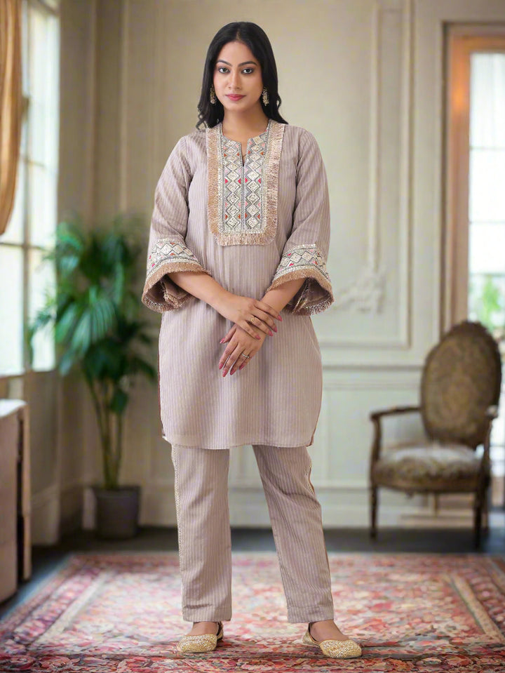 Woman wearing a beige embroidered kurta with matching pants against a white paneled wall.