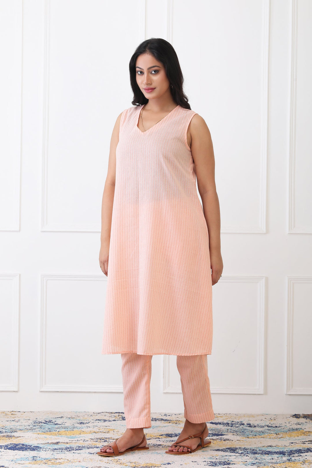 Woman wearing a peach sleeveless top and pants against a white paneled wall.