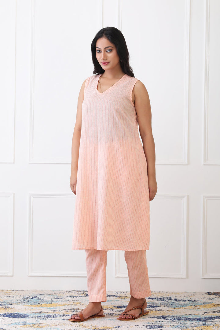 Woman wearing a peach sleeveless top and pants against a white paneled wall.