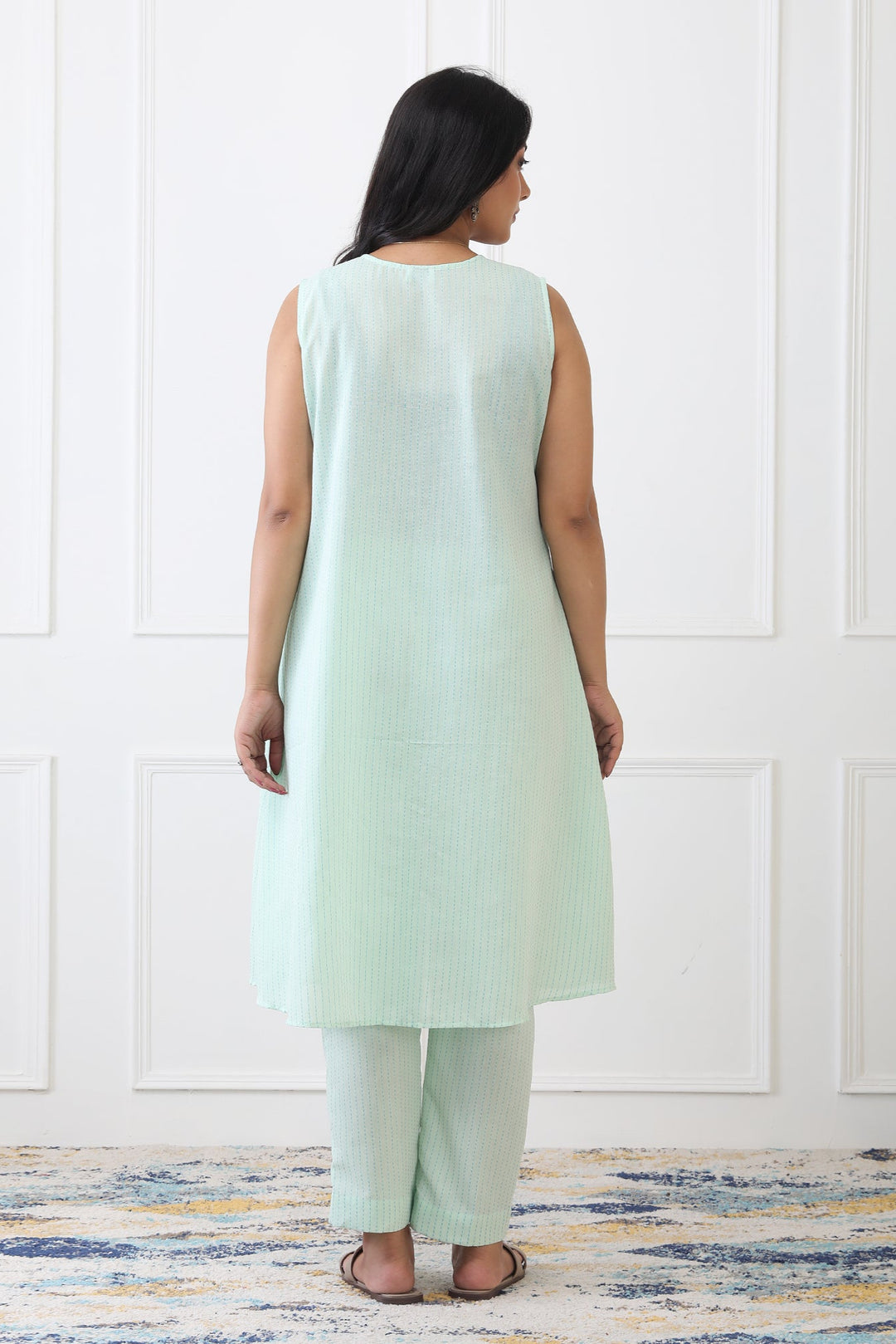 Woman wearing a light green sleeveless top and pants against a white paneled wall.