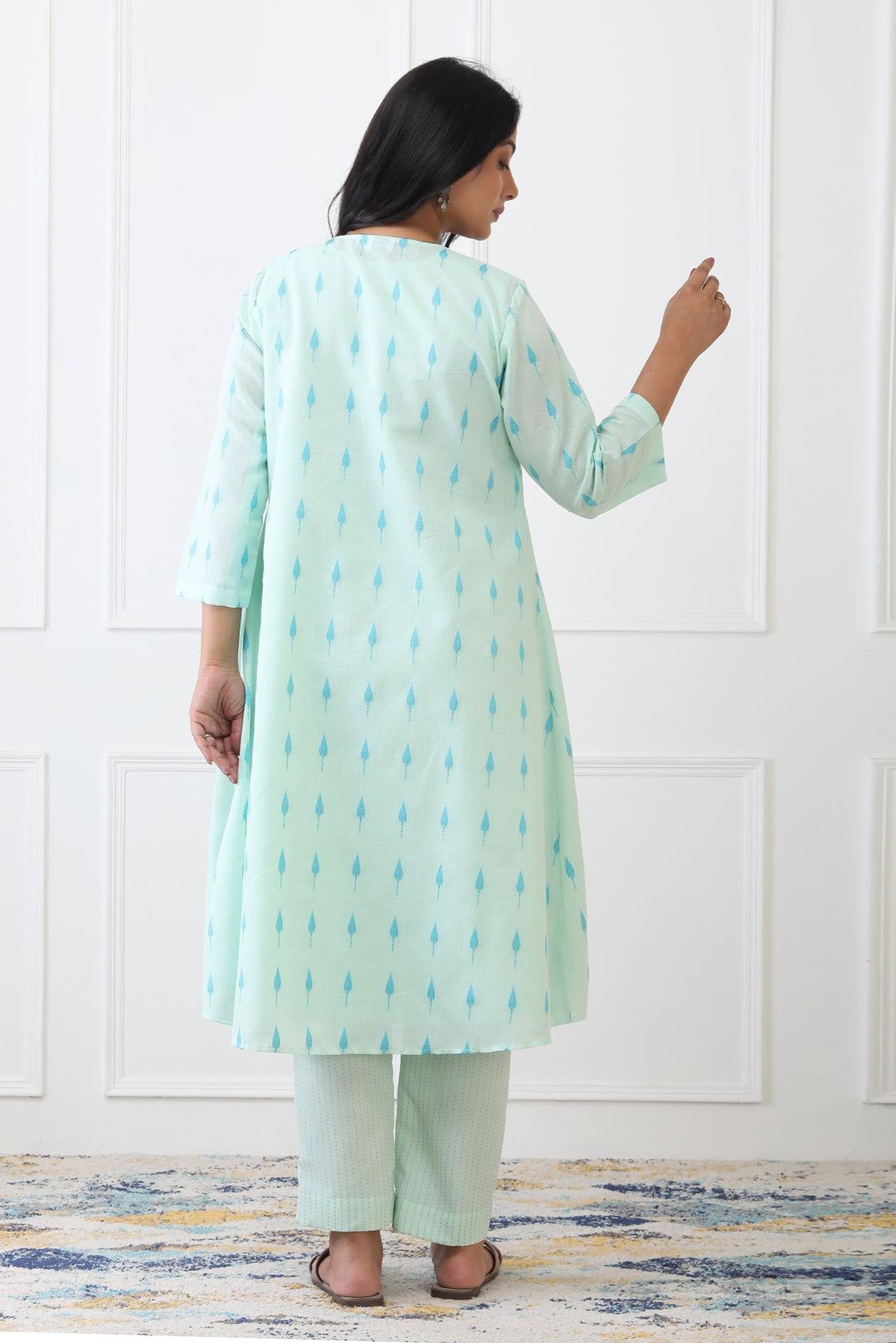 Woman wearing a light green kurta with white pants against a white paneled wall.