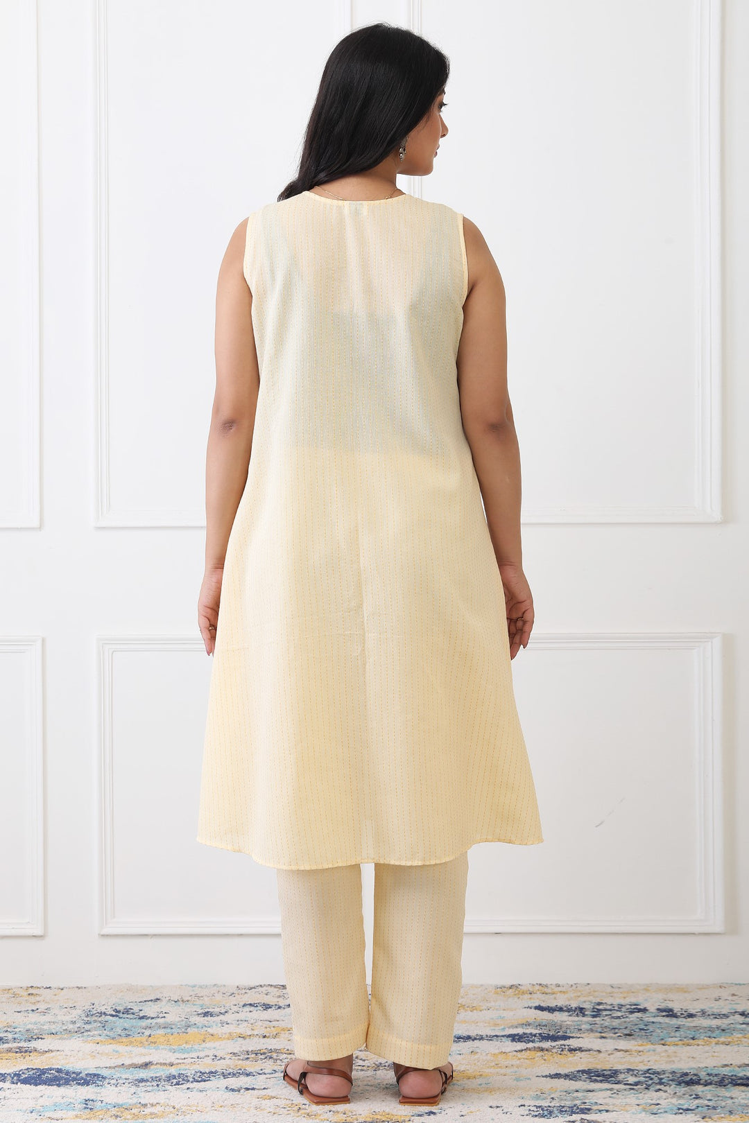 Woman wearing a light yellow sleeveless top and pants against a white wall.