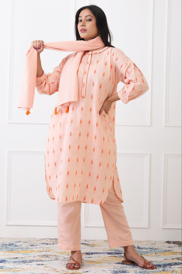 Woman wearing a peach-colored outfit with a matching scarf against a white paneled wall.