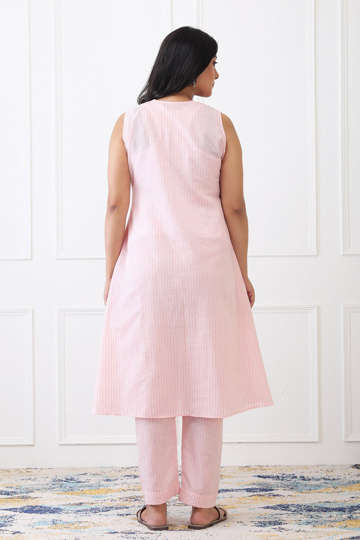Woman wearing a light pink sleeveless top and pants against a white paneled wall.