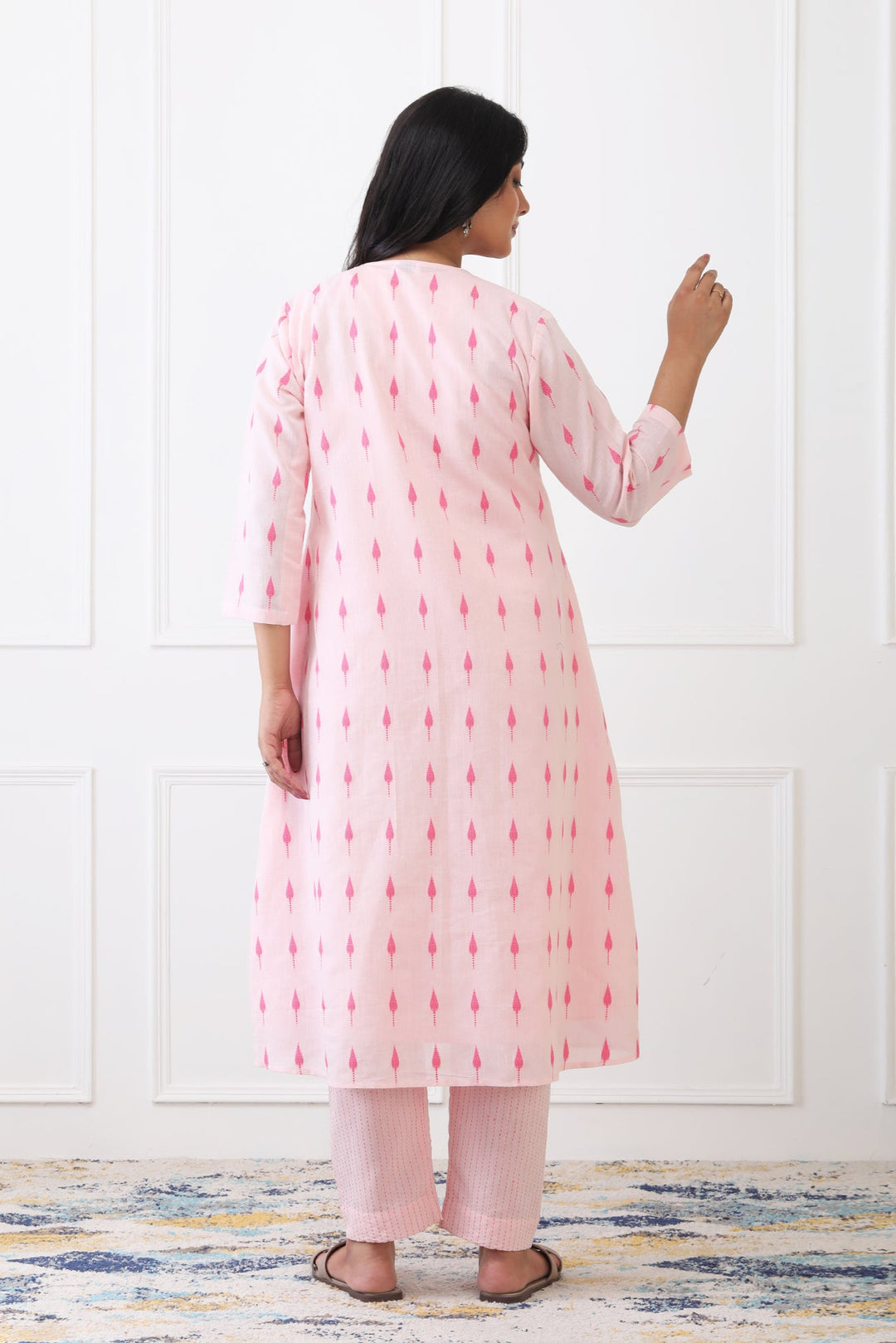 Woman wearing a pink kurta with red patterns in a room with a white paneled wall.