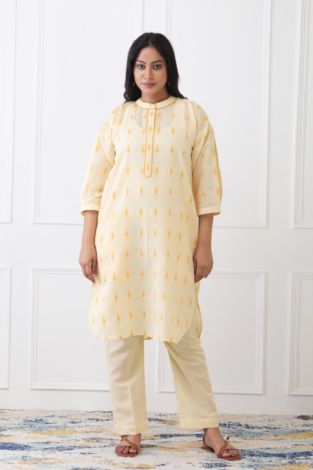 Woman wearing a light yellow kurta with white pants against a white paneled wall.
