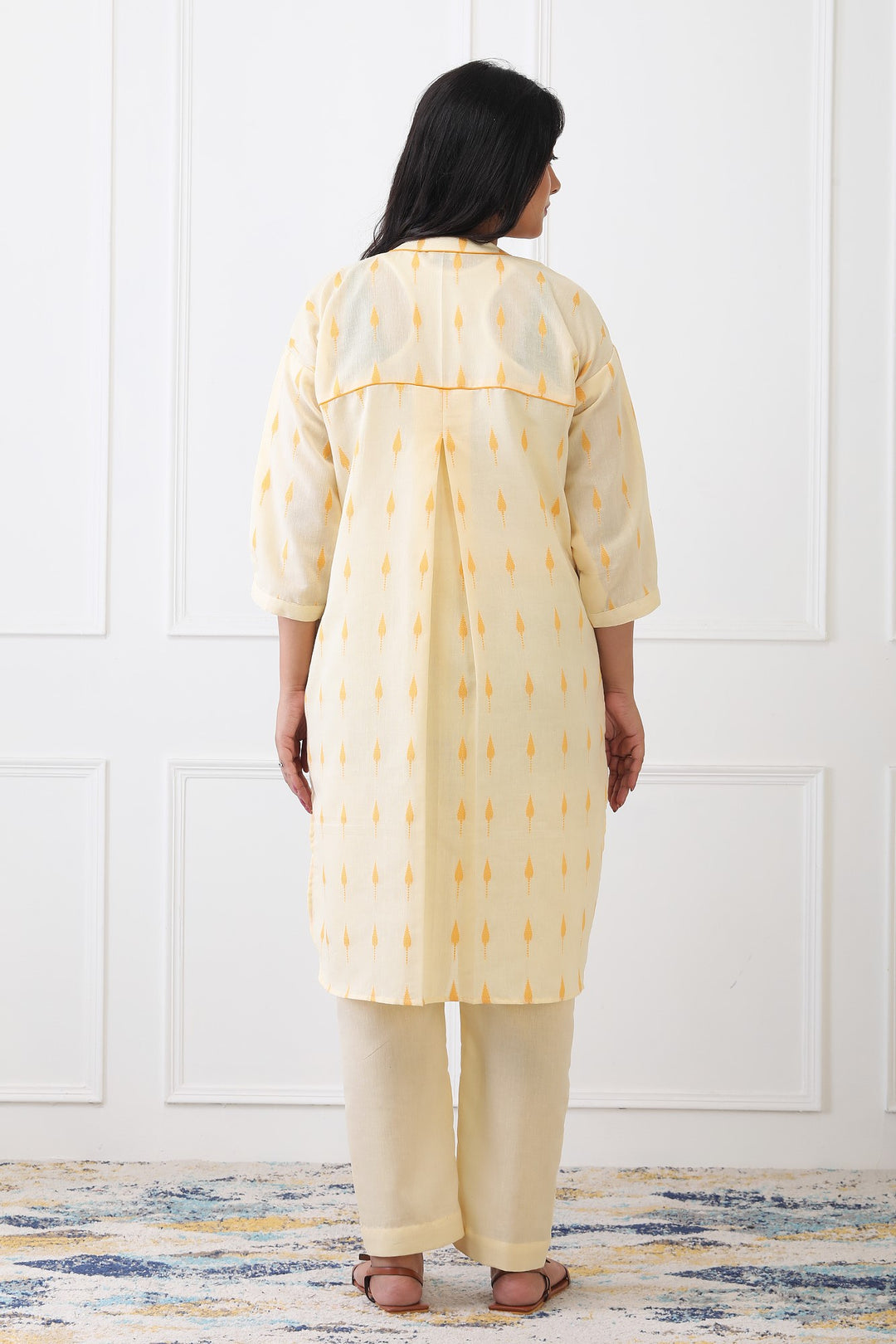 Woman wearing a yellow kurta with white patterns against a white paneled wall.