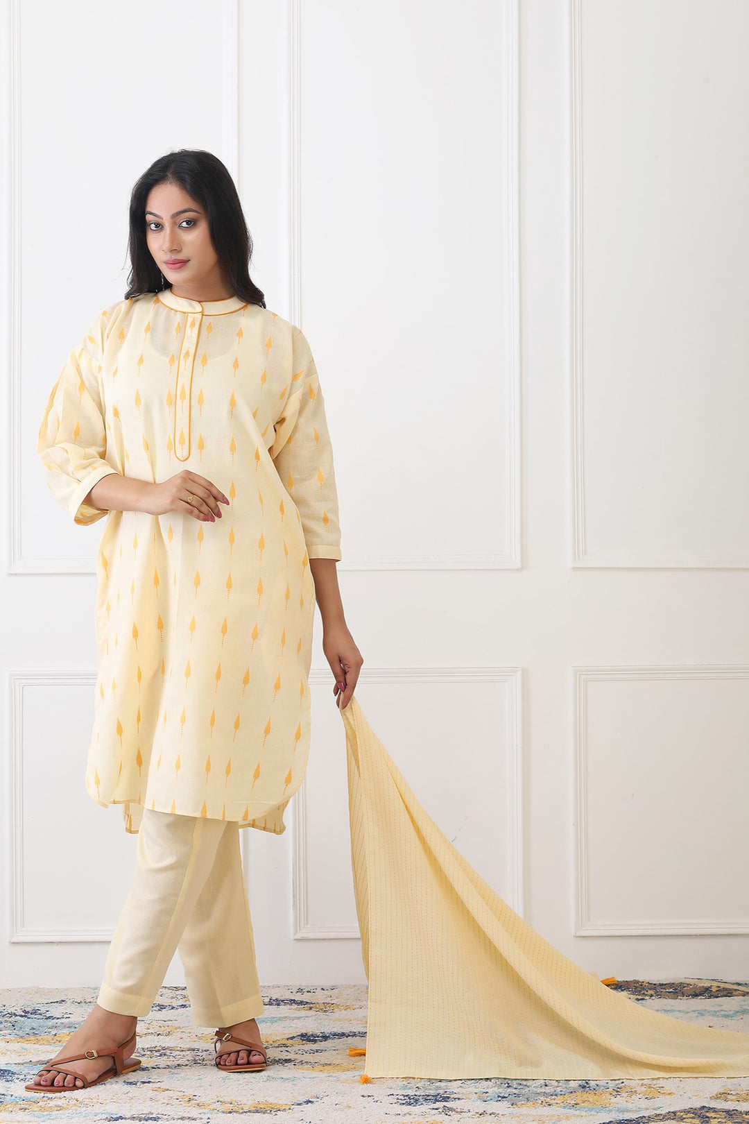 Woman wearing a light yellow kurta with white pants, holding a matching dupatta against a white wall.