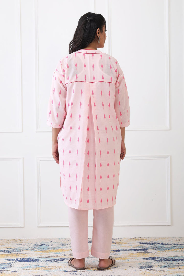 Woman wearing a pink patterned kurta with white pants against a white paneled wall.