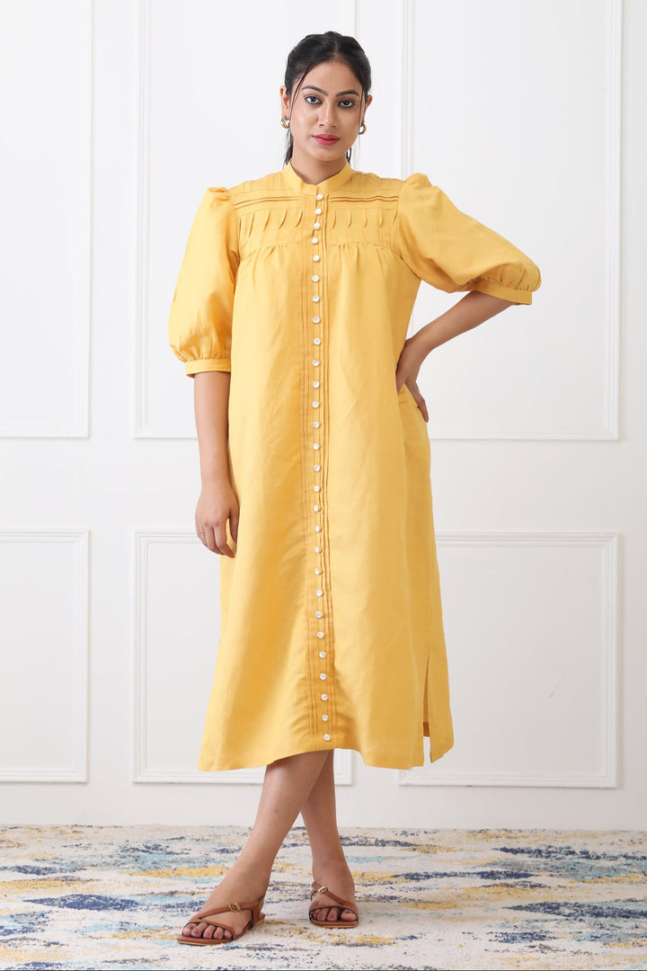 Woman wearing a yellow dress with puff sleeves against a white wall.