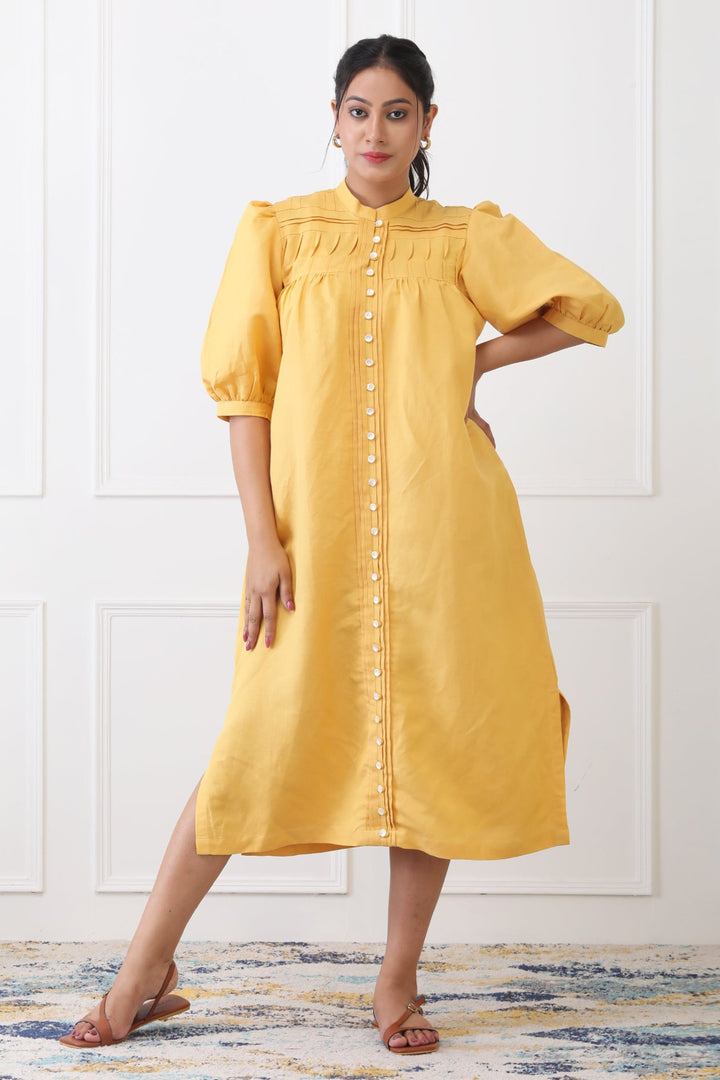 Woman wearing a yellow dress with puff sleeves against a white wall.