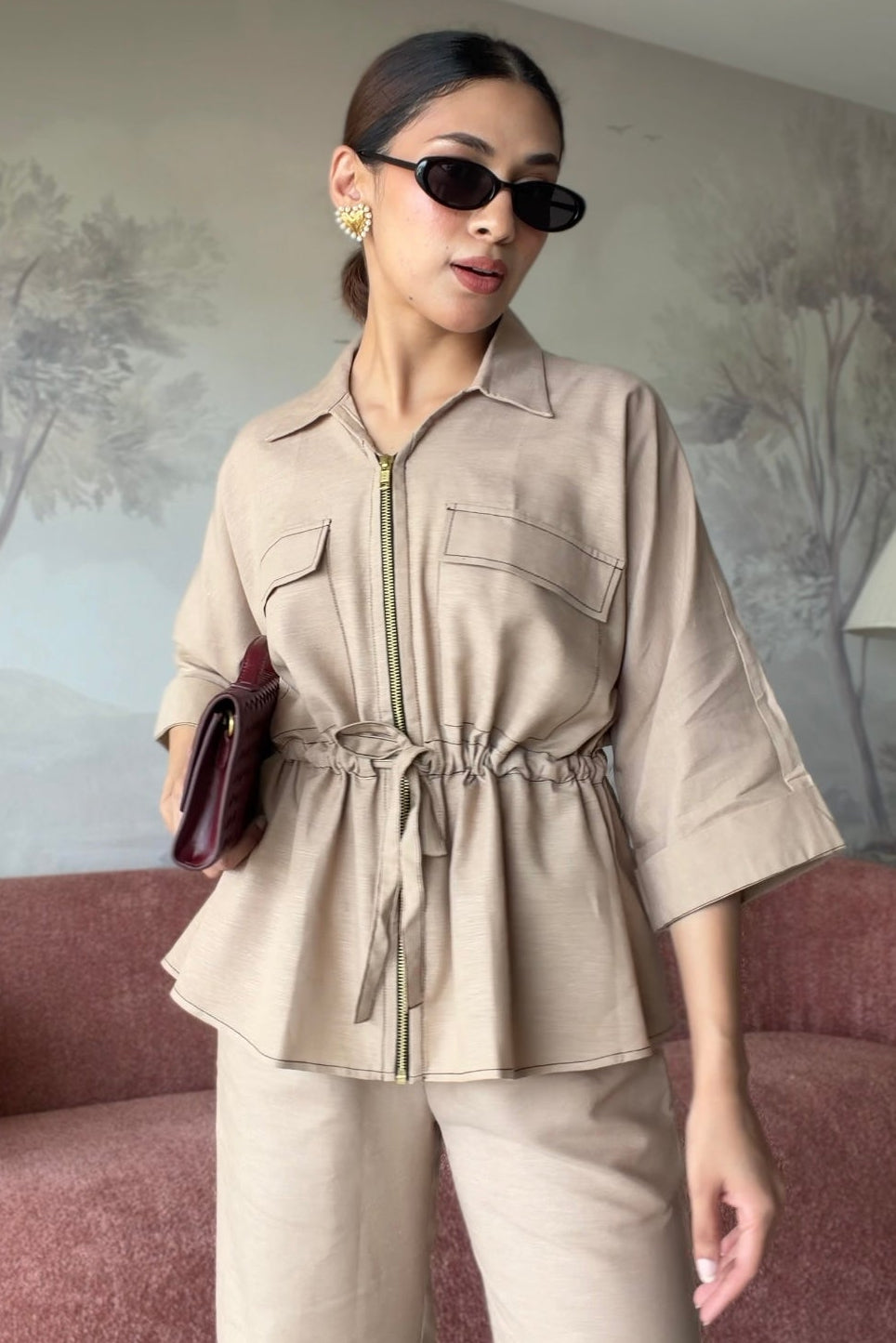A woman models a beige two-piece jumpsuit set consisting of a collared zip-front top with a drawstring waist and matching wide-leg trousers, styled in a living room setting with a pink sofa and tree-patterned wallpaper.
