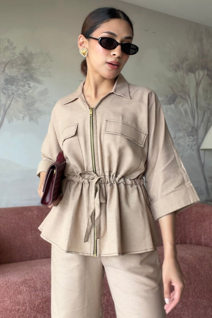 A woman models a beige two-piece jumpsuit set consisting of a collared zip-front top with a drawstring waist and matching wide-leg trousers, styled in a living room setting with a pink sofa and tree-patterned wallpaper.