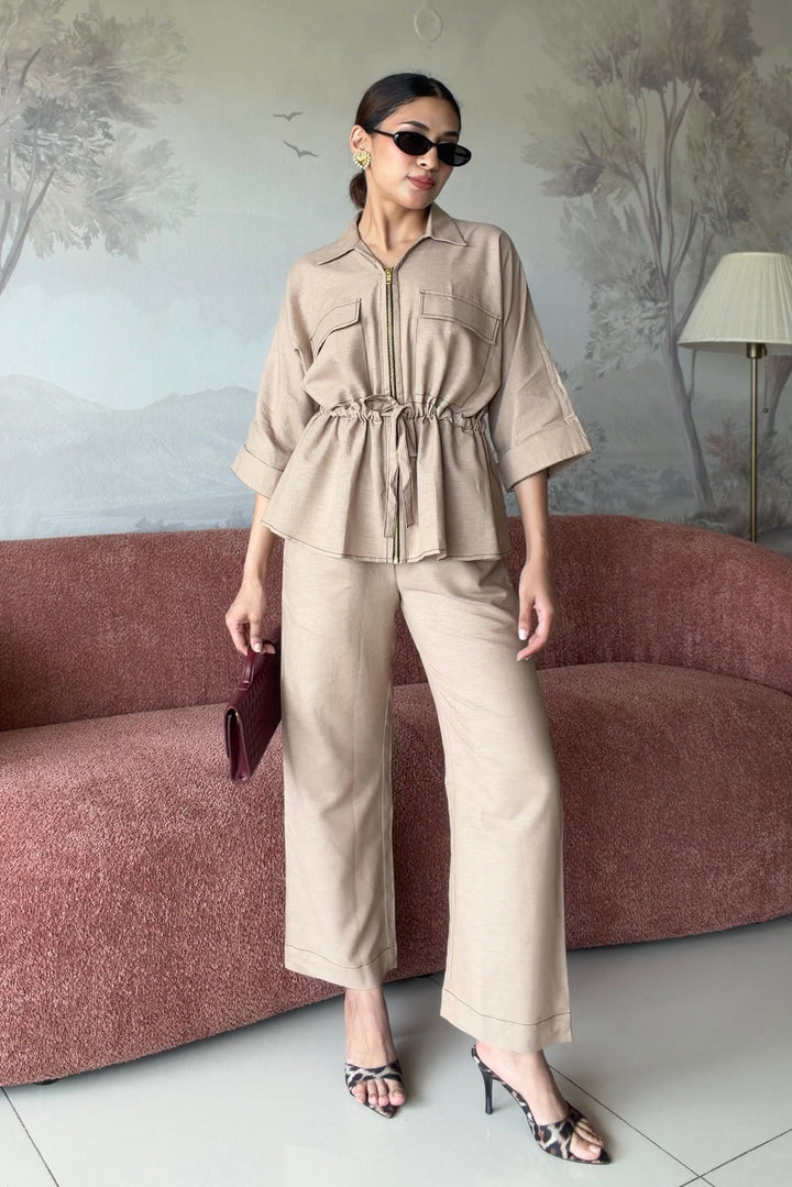 A woman models a beige two-piece matching set consisting of a short-sleeve button-up shirt with a drawstring waist and matching wide-leg trousers, styled with black sunglasses, a dark burgundy clutch, and black heeled sandals in front of a dusty rose textured sofa.