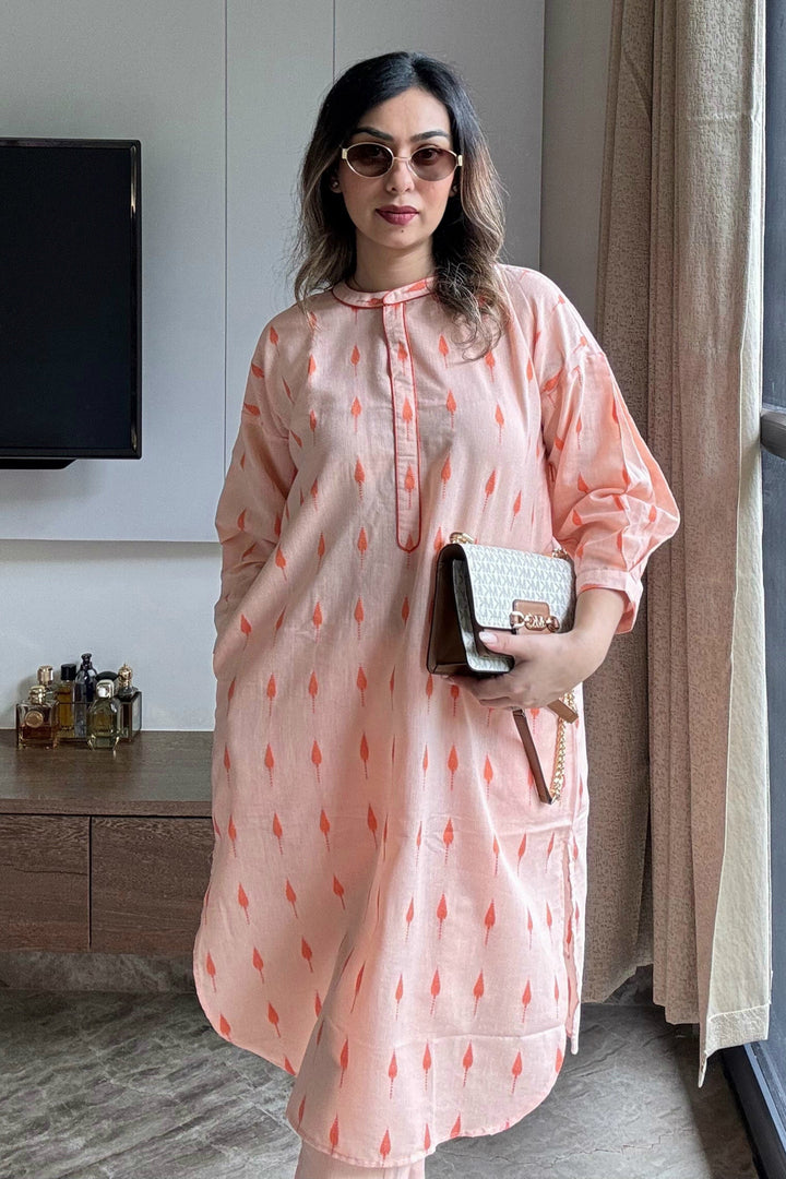 Woman in a peach-colored dress with red patterns standing in a room with a television and window.