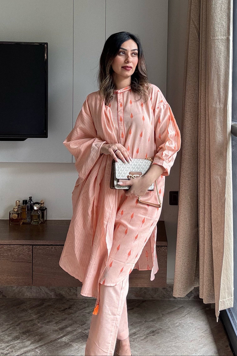 Woman in a peach outfit standing in a room with a curtain and television.