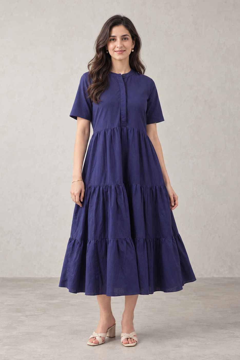 A woman modeling a short-sleeve, tiered midi dress in a deep navy blue color, standing against a textured light gray wall.