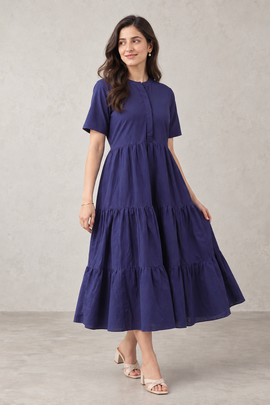 A woman modeling a short-sleeve midi dress in deep navy blue, featuring a tiered skirt and a buttoned round neckline, set against a textured light gray studio backdrop.