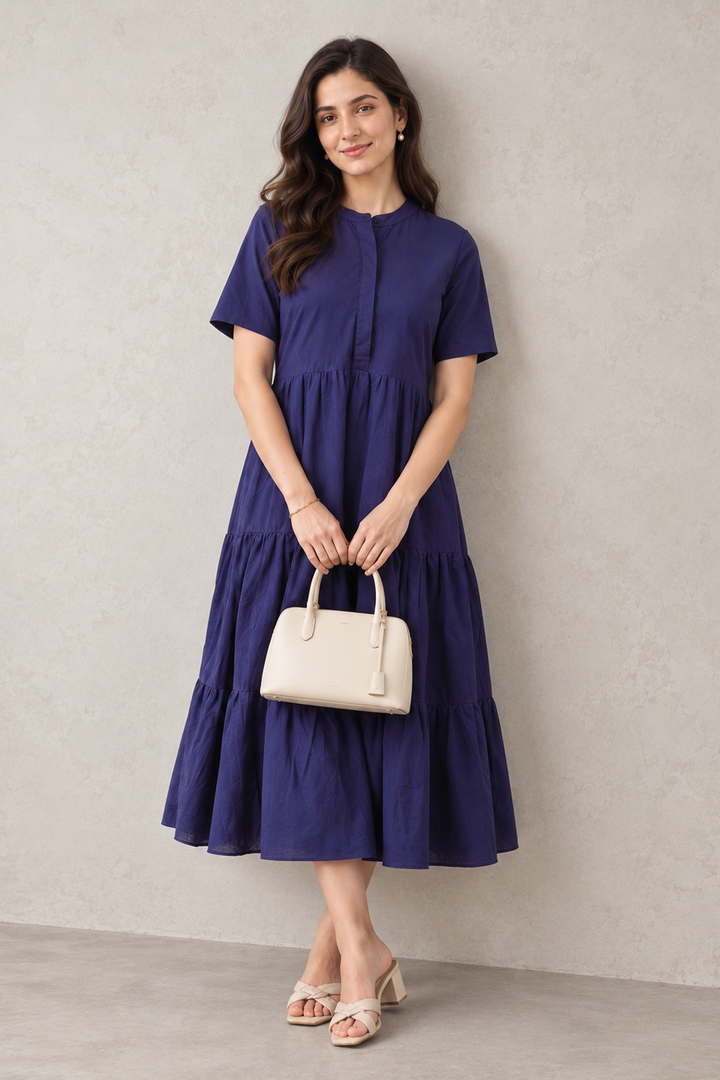 A woman stands against a textured light gray wall wearing a short-sleeve tiered midi dress in deep navy blue. She is holding a cream-colored handbag and wearing beige heeled sandals.