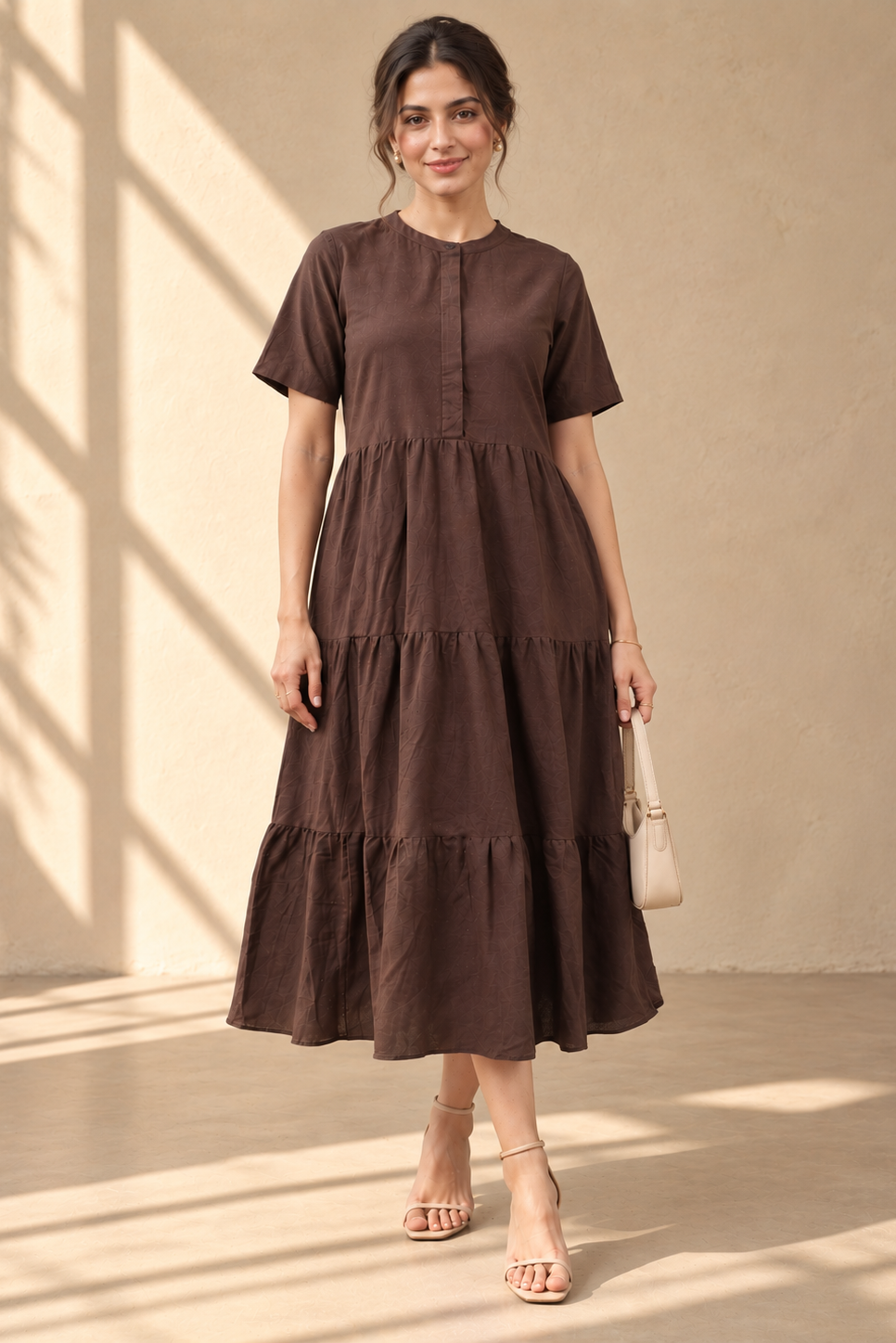 A woman models a dark brown tiered midi dress with short sleeves and a buttoned neckline, standing in a sunlit room with warm beige walls and floor shadows.