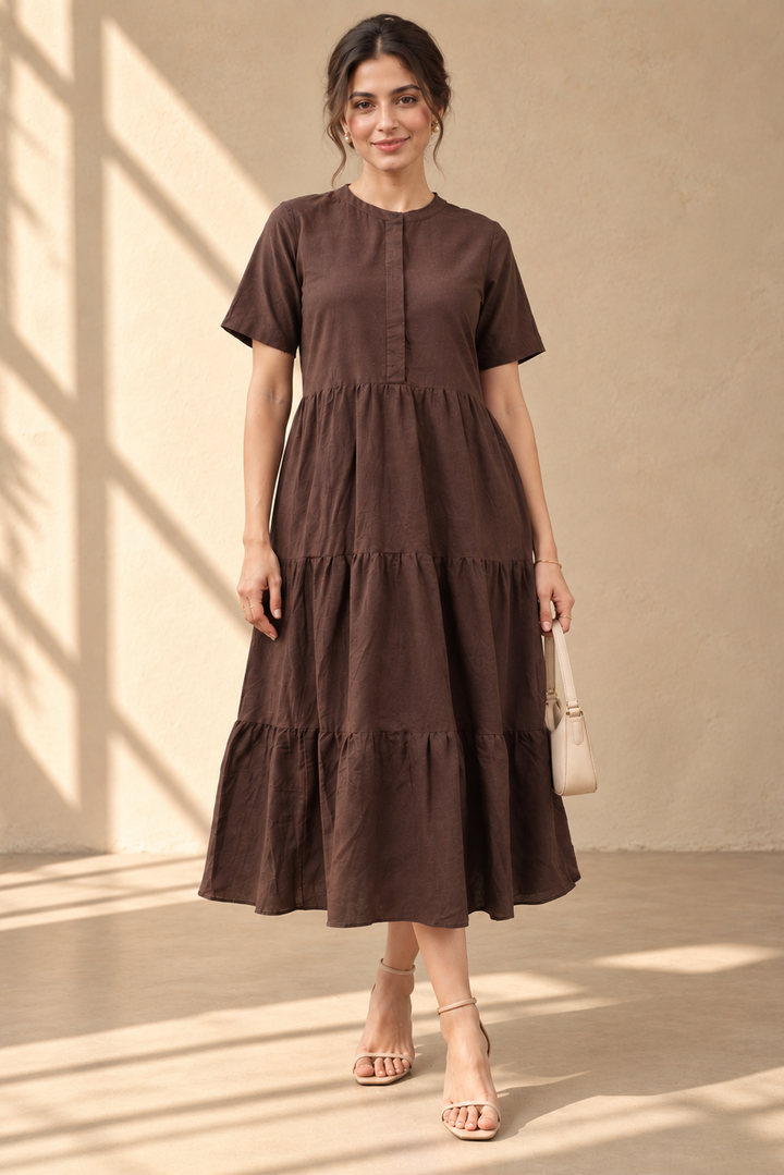 A woman models a dark brown tiered midi dress with short sleeves and a buttoned neckline, standing in a sunlit room with warm beige walls and floor shadows.