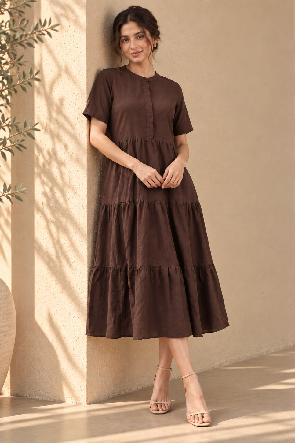 A woman wearing a dark brown tiered midi dress with short sleeves and a buttoned round neckline, standing against a beige stucco wall with warm sunlight and shadows.