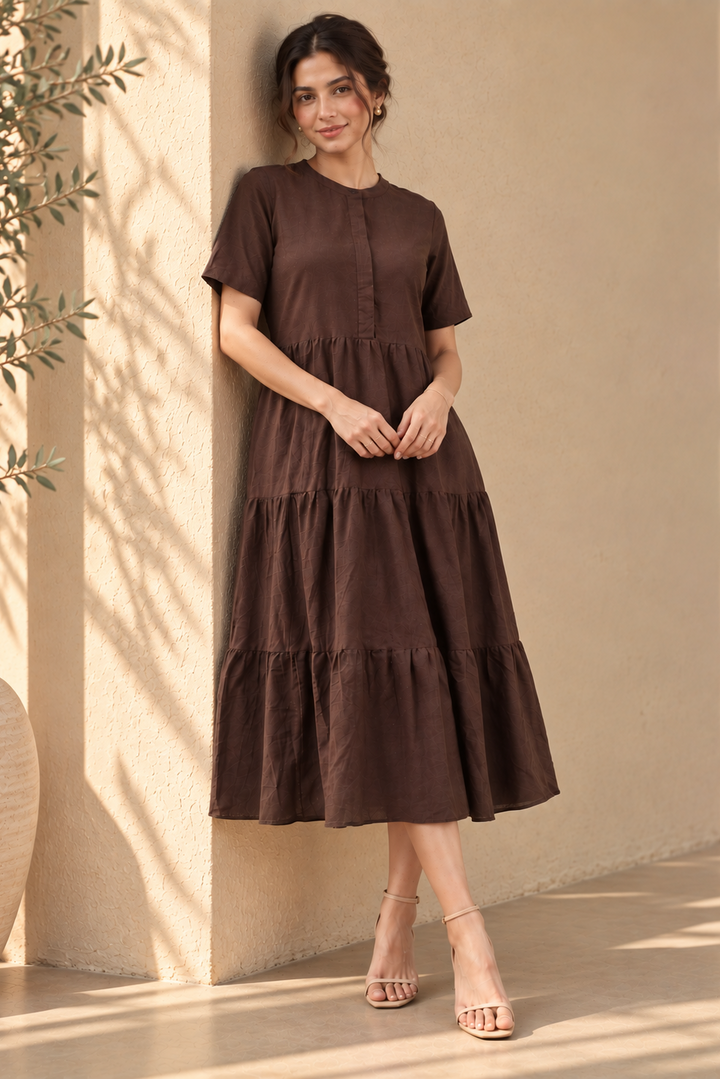 A woman wearing a dark brown tiered midi dress with short sleeves and a buttoned round neckline, standing against a beige stucco wall with warm sunlight and shadows.